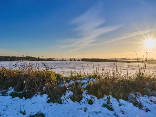 This landscape photograph captures the rural fields near Bolsover, covered in a layer of snow during the winter season. The sun is low on the horizon, casting a warm glow across the snow-covered fields and creating long shadows in the foreground. With clear skies and some wispy clouds, the image reflects a peaceful winter afternoon in the countryside, highlighting the natural beauty of Bolsover’s rural landscape at this time of year.